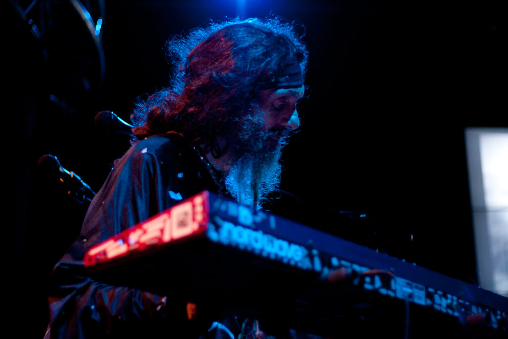 Mężczyzna w długich włosach i z brodą w oświetleniu czerwono-niebieskim siedzi przed keyboardem