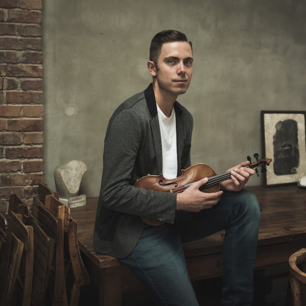 Mateusz Smoczyński siedzi na stole, w rękach trzyma skrzypce