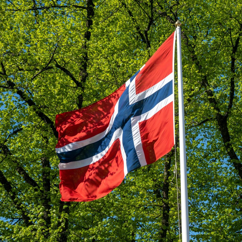czerwono granatowa flaga Norwegii powiewa na maszcie na tle drzew z liścmi