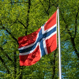 czerwono granatowa flaga Norwegii powiewa na maszcie na tle drzew z liścmi