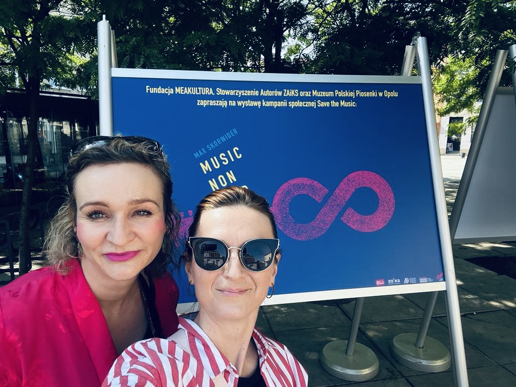 dwie uśmiechnięte kobiety robią selfie na tle granatowego plakatu na wystawie MUSIC NON STOP