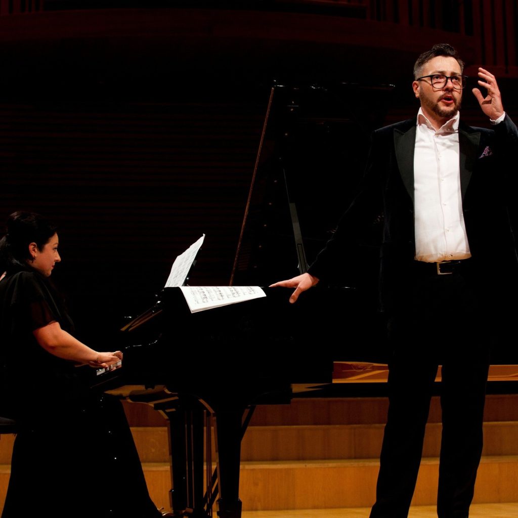 Zdjęcie przedstawia wokalistę Jakuba Schmidta, podczas występu na scenie z towarzyszeniem fortepianu, przy którym zasiada pianistka Mirella Malorny.