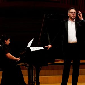 Zdjęcie przedstawia wokalistę Jakuba Schmidta, podczas występu na scenie z towarzyszeniem fortepianu, przy którym zasiada pianistka Mirella Malorny.