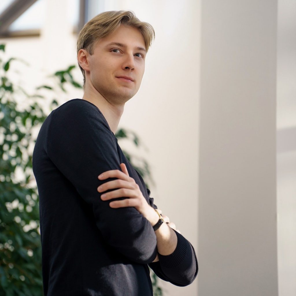 Portret Juliana Gilewskiego - młodego dyrygenta - na tle białego budynku i zielonej rośliny