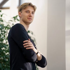 Portret Juliana Gilewskiego - młodego dyrygenta - na tle białego budynku i zielonej rośliny